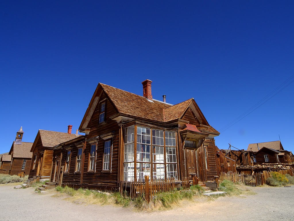 A weathered timber house with a glazed porch and uneven fence in Bodie, beneath a deep blue sky.