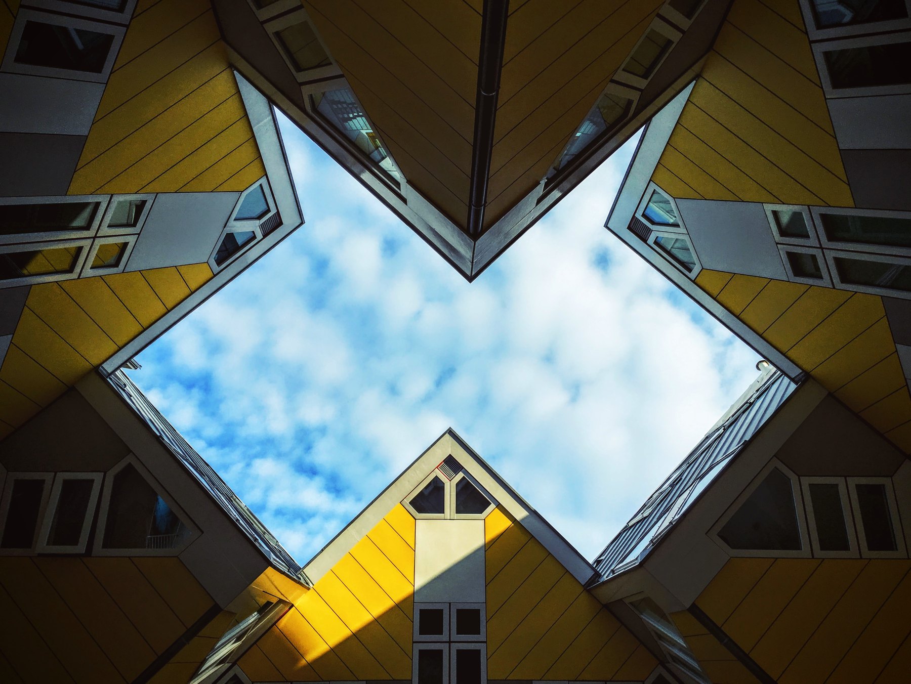 Looking straight up between yellow Cube Houses, with sloping window bands framing a diamond-shaped patch of blue sky.