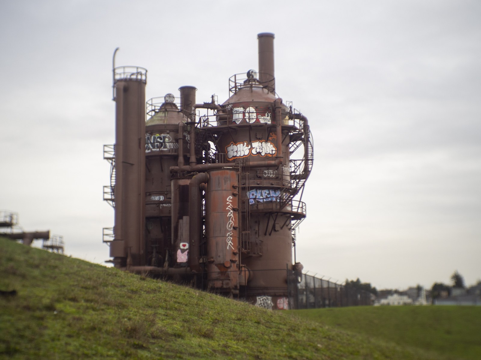 Rust-coloured gas-processing towers covered in graffiti rise behind a fence and grassy slopes at Gas Works Park.