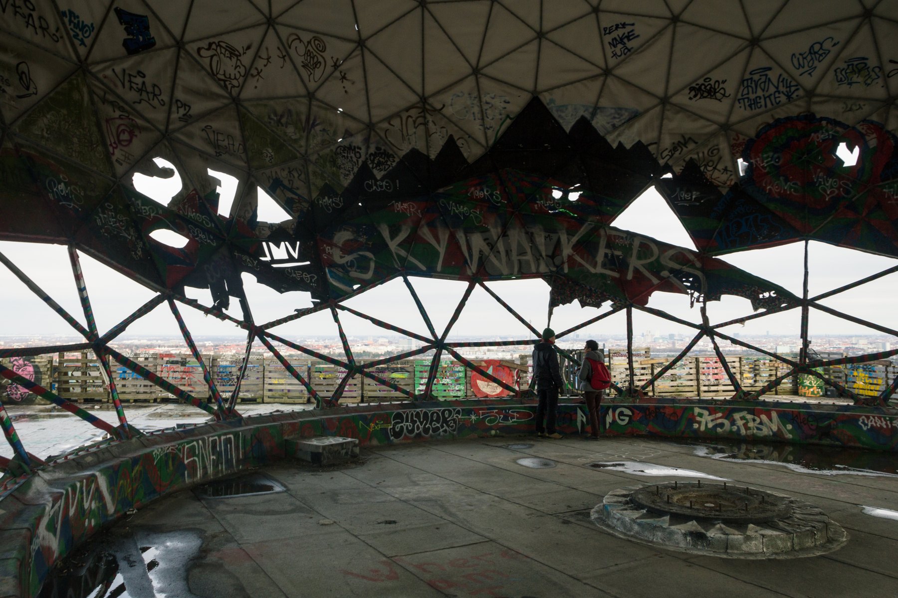 Inside a damaged dome at Teufelsberg, triangular steel framing opens onto a view of Berlin beyond graffiti-covered walls.