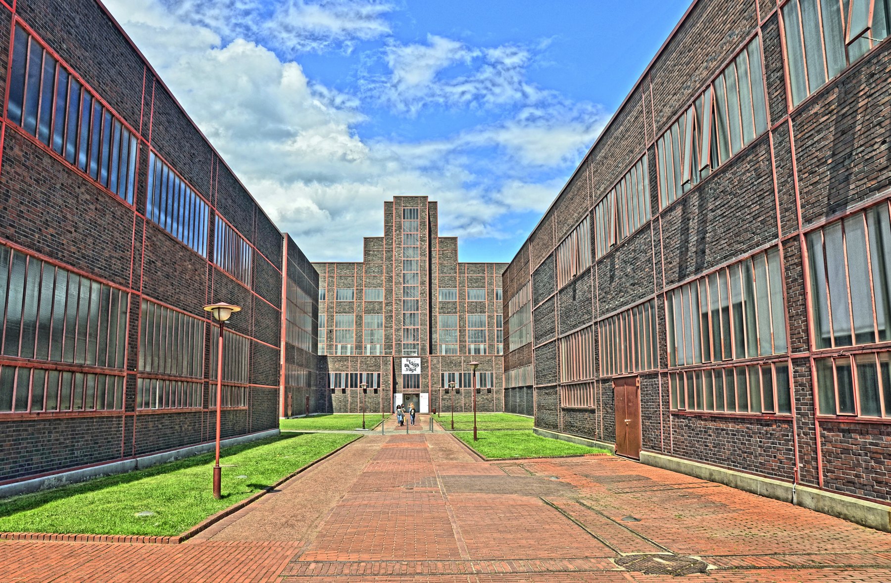 Brick path between long red-framed industrial buildings at Zollverein, with a tall central window ahead.